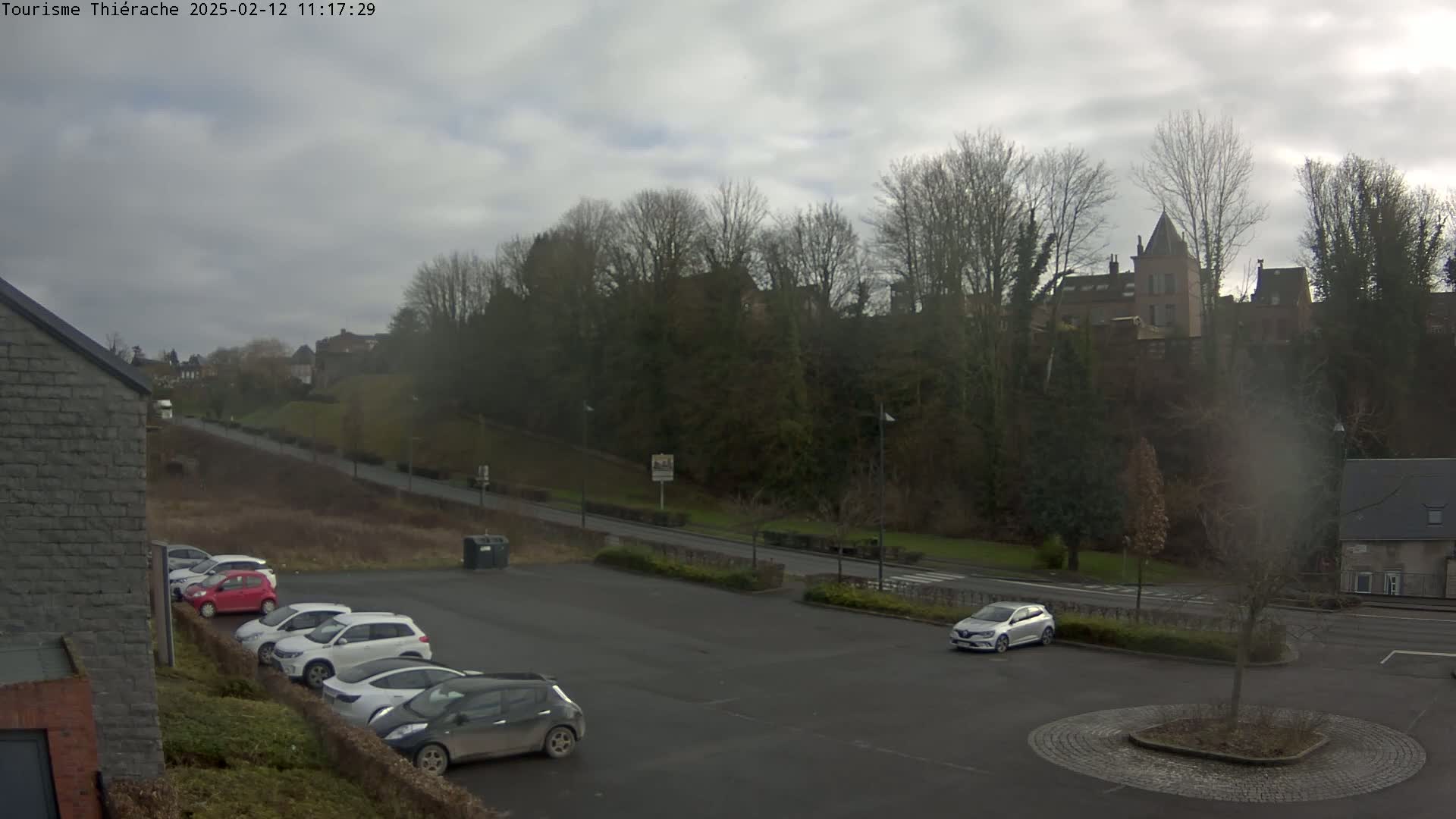 A parking lot with several cars is situated below a hill on which buildings and trees are visible under an overcast sky.