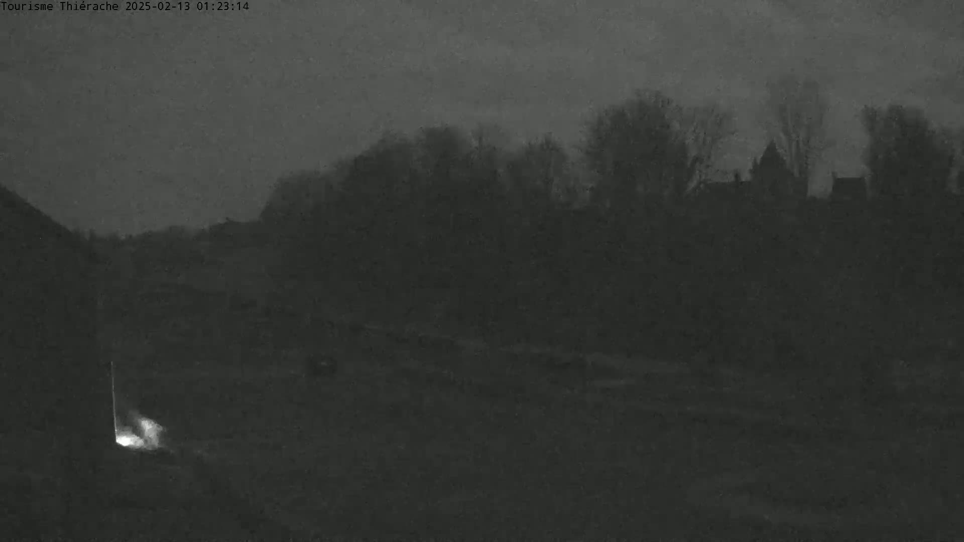 A dimly lit, nighttime scene shows a faint light near the bottom left corner and dark silhouettes of trees and buildings in the distance under a dark, overcast sky.