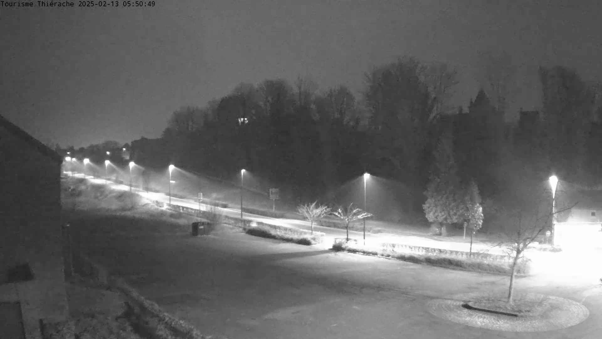 A snow-covered road at night, illuminated by streetlights, winds past a line of trees towards a building in the distance.