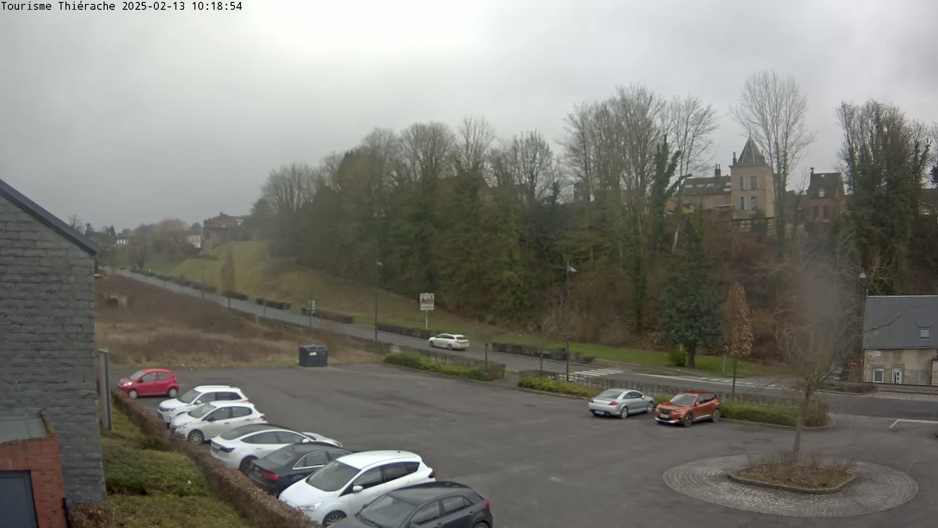 A parking lot with several cars is situated below a hill covered in trees and a building, under an overcast sky.