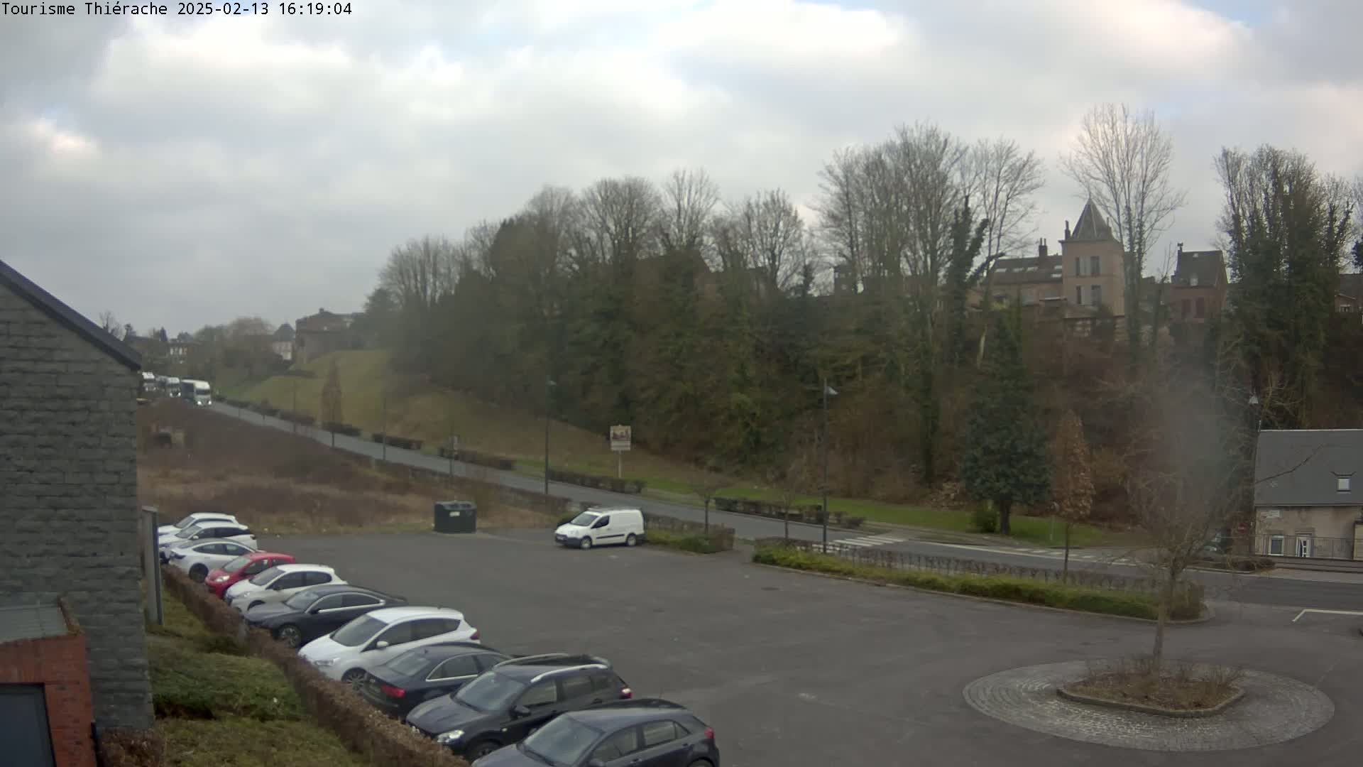A parking lot with several cars is situated below a hill with trees and a building, under a cloudy sky.