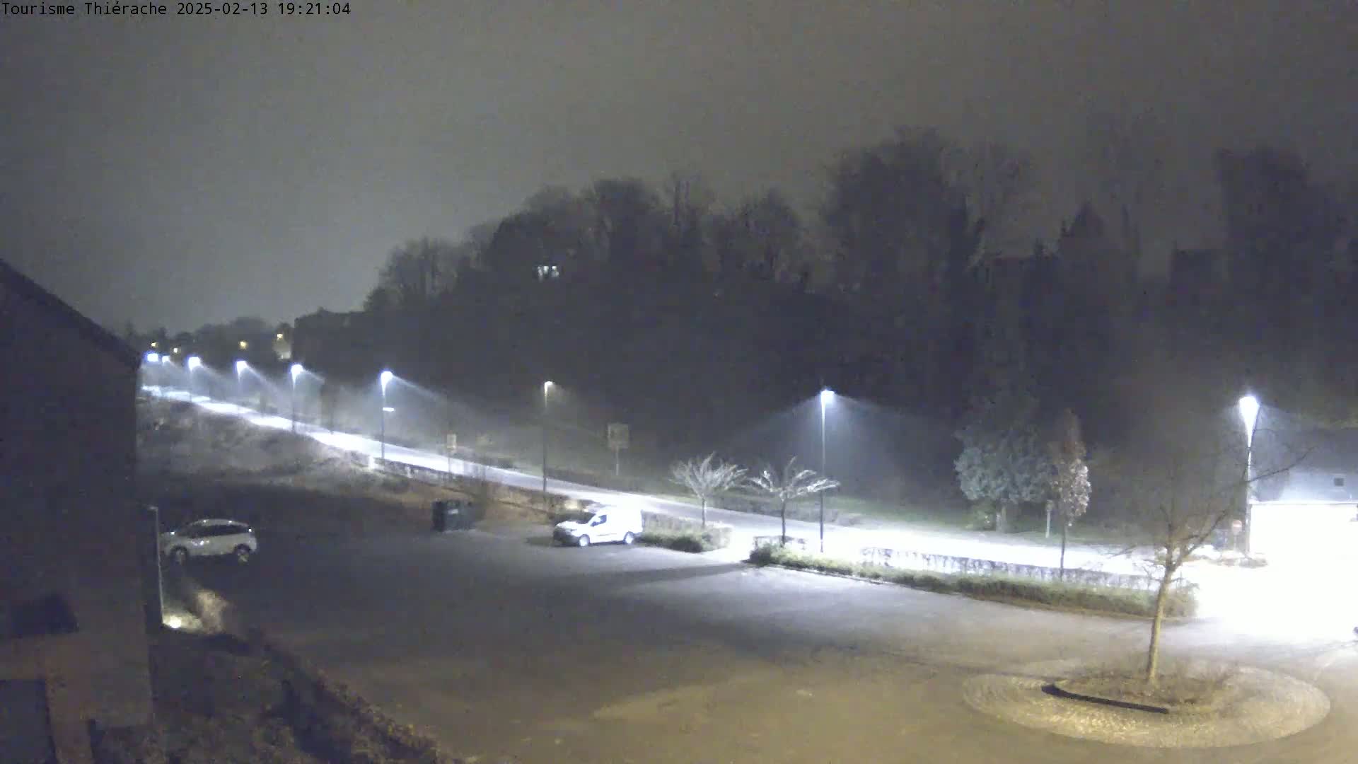 A nighttime, foggy scene shows a mostly empty parking lot with a few cars, illuminated by streetlights, and a road leading to a dark, tree-covered hill in the background.