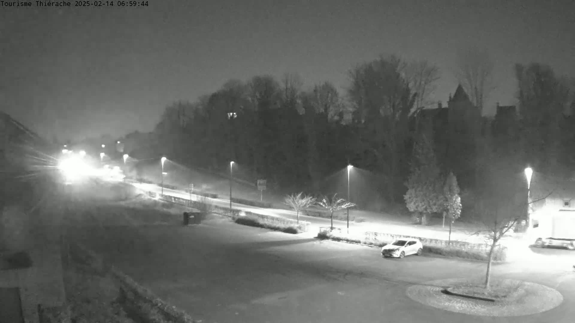 A nighttime, snow-covered parking lot and road are illuminated by streetlights, with a few vehicles visible and a line of trees and buildings in the background.