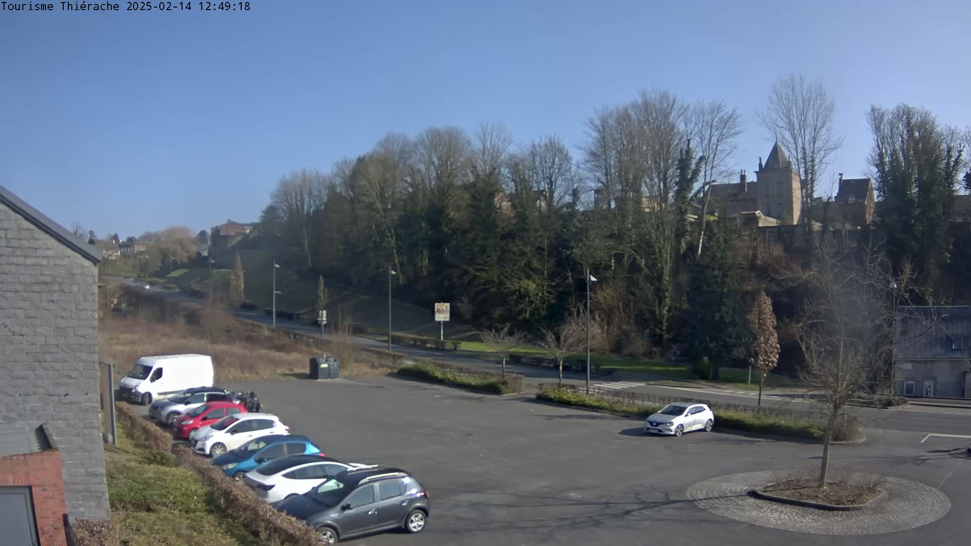 A parking lot with several cars is situated below a hill covered in leafless trees and buildings under a clear, sunny sky.