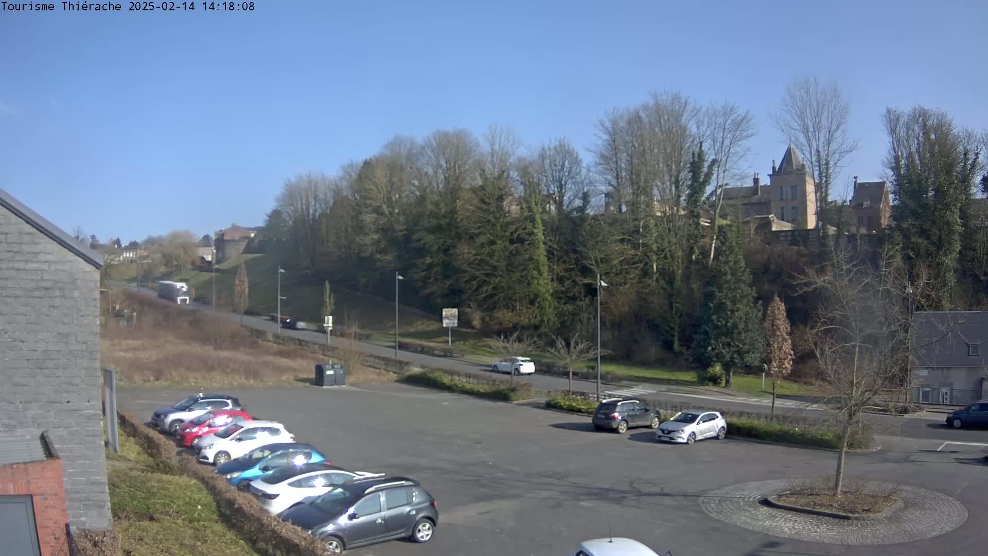 A parking lot with several cars is situated in front of a roadway leading to a hill covered with trees and topped with a castle-like structure, all under a clear sunny sky.