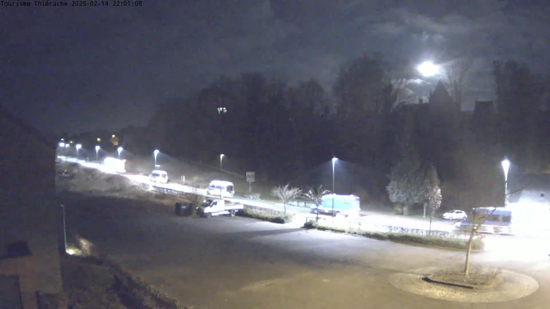 Several vehicles are parked and driving on a road at night under a partly cloudy sky illuminated by a bright moon.
