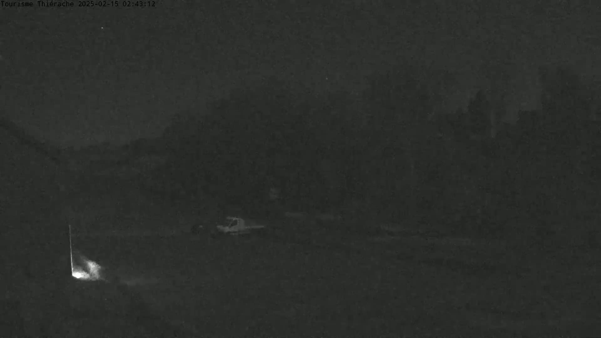 A low-light, nighttime image shows a dark, mostly empty landscape with a few indistinct objects and a bright light source near the bottom left.