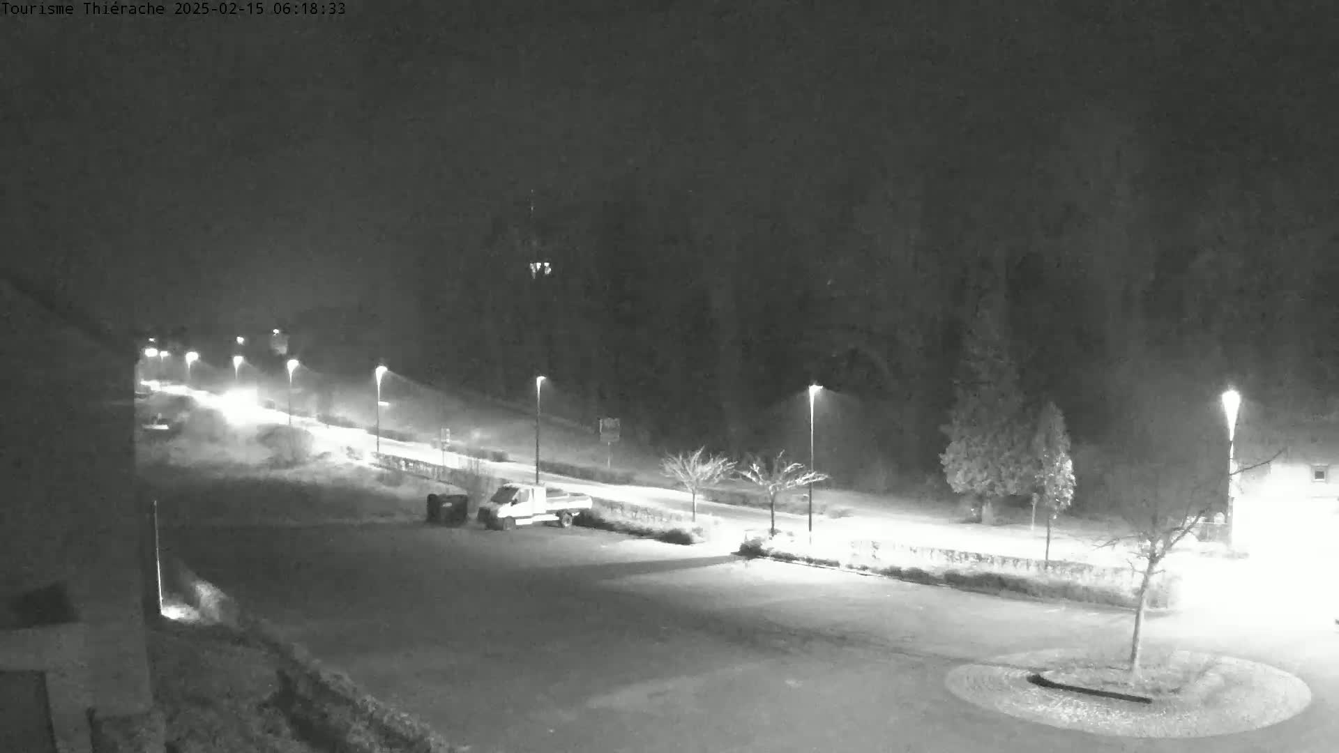 A nighttime, snowy scene shows a road with cars and streetlights, a parking lot with a van, and bare trees in the background.