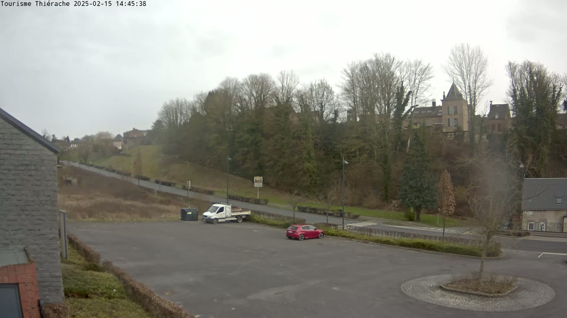 An overcast day reveals a parking lot with a white van and a red car, overlooking a road that leads to a hill topped with buildings and trees.
