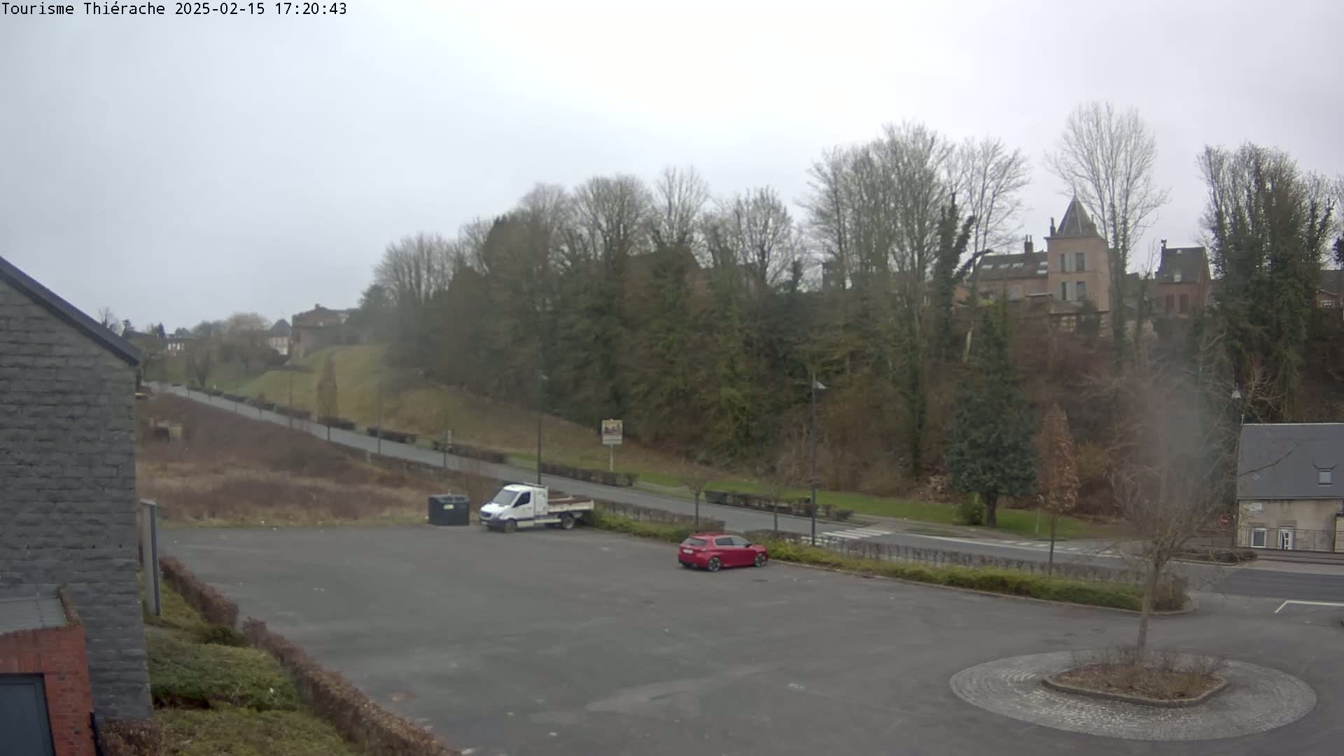 An overcast day reveals a mostly empty parking lot with a white van and a red car, overlooking a tree-lined hill with buildings in the distance.