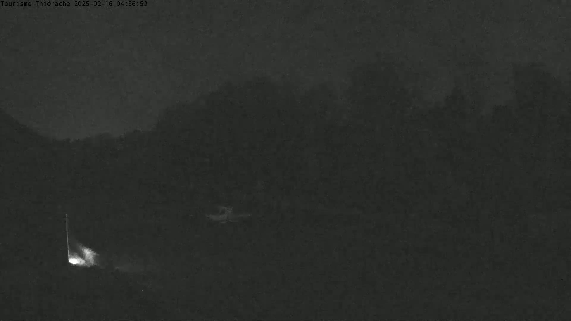 A nighttime, low-light image shows a dark, indistinct landscape with a small, bright light source near the bottom left.
