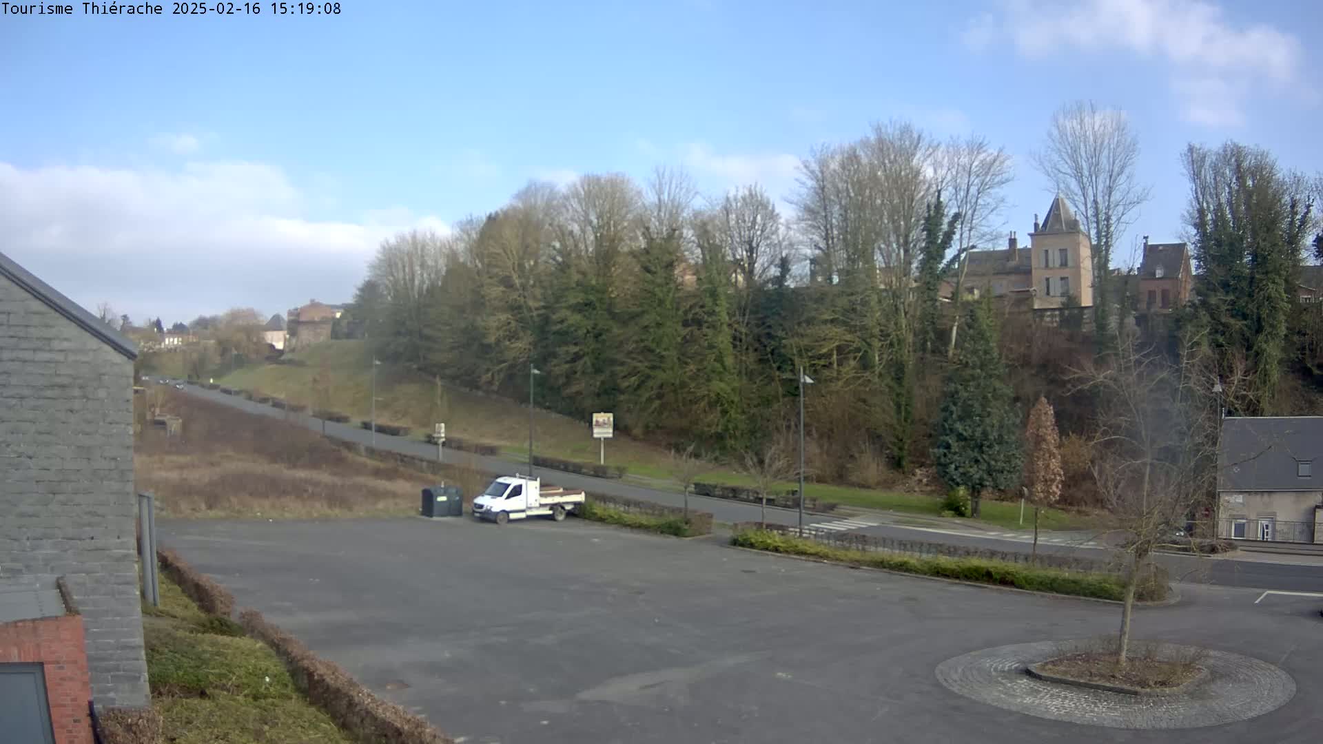 A white pickup truck is parked in an empty lot overlooking a road that leads to a hill covered in leafless trees and buildings under a mostly clear, sunny sky.