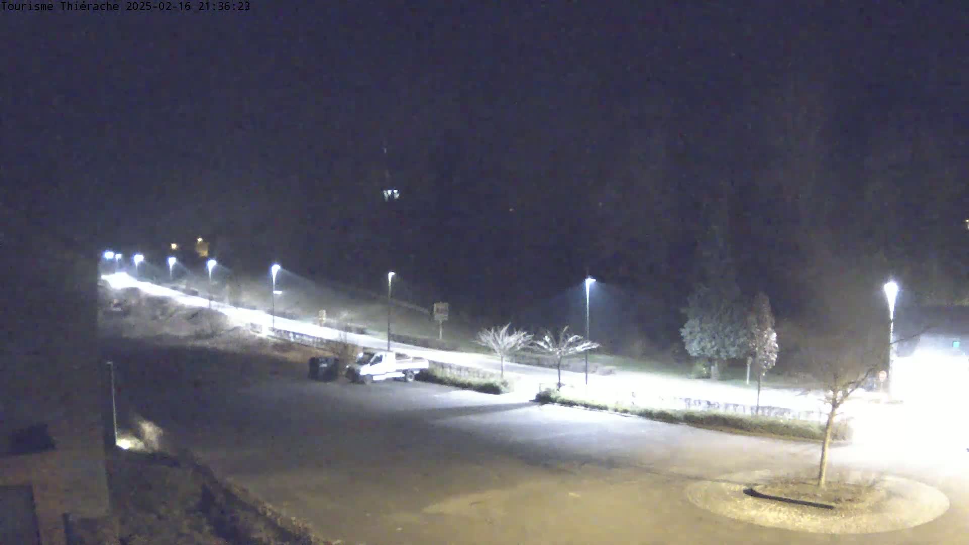 A nighttime view of a mostly empty parking lot and a road lined with streetlights, under a dark, possibly foggy sky.