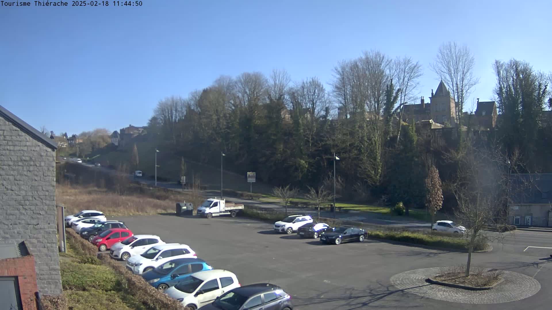 A sunny day shows a parking lot filled with cars in front of a tree-covered hill with buildings on top.
