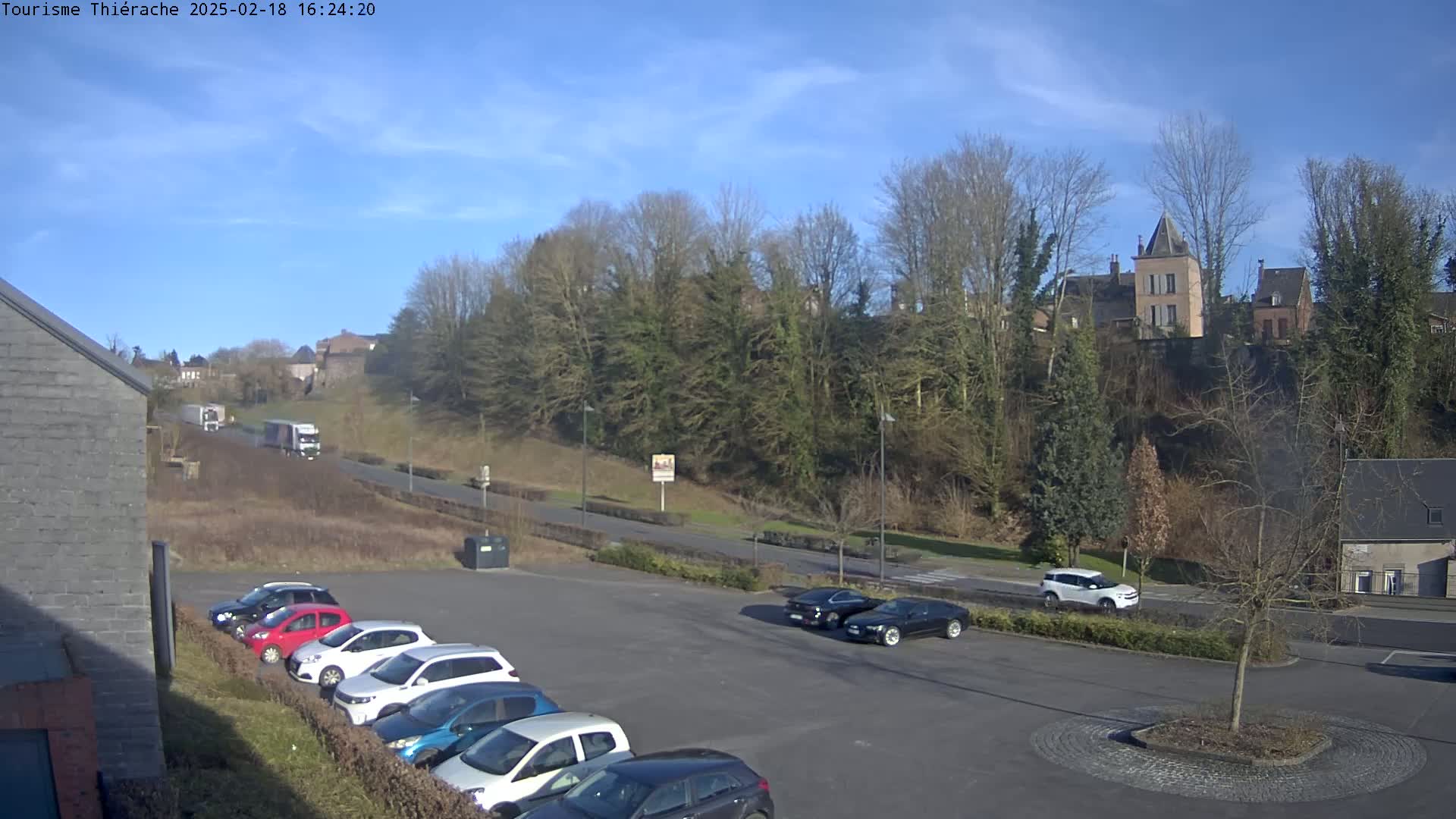 A parking lot with several cars is situated near a road, beyond which lies a hill covered in leafless trees and topped with buildings under a clear, sunny sky.