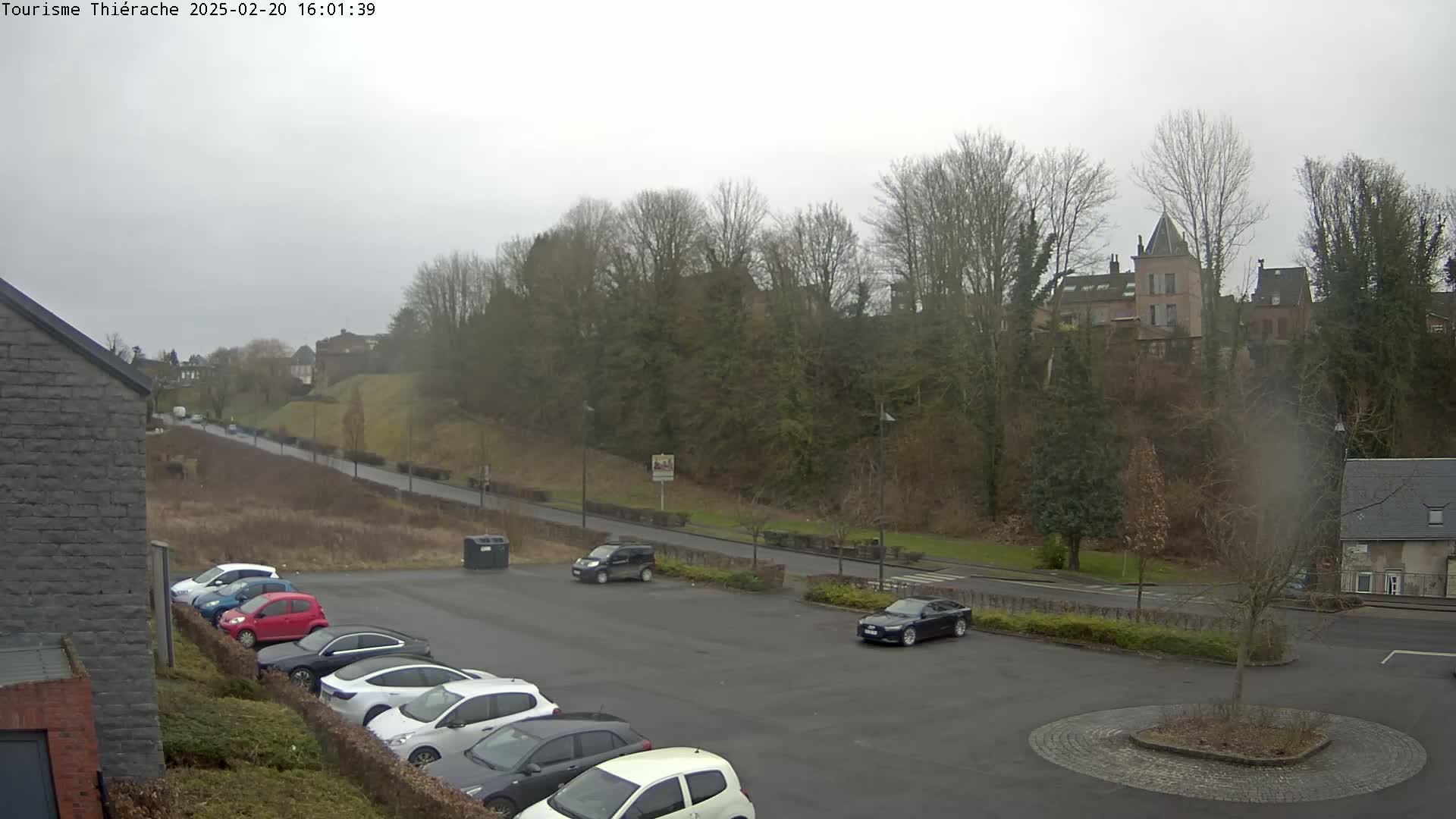 A parking lot with several cars is situated in front of a hill covered in bare winter trees, overlooking a building with a tall tower under an overcast sky.
