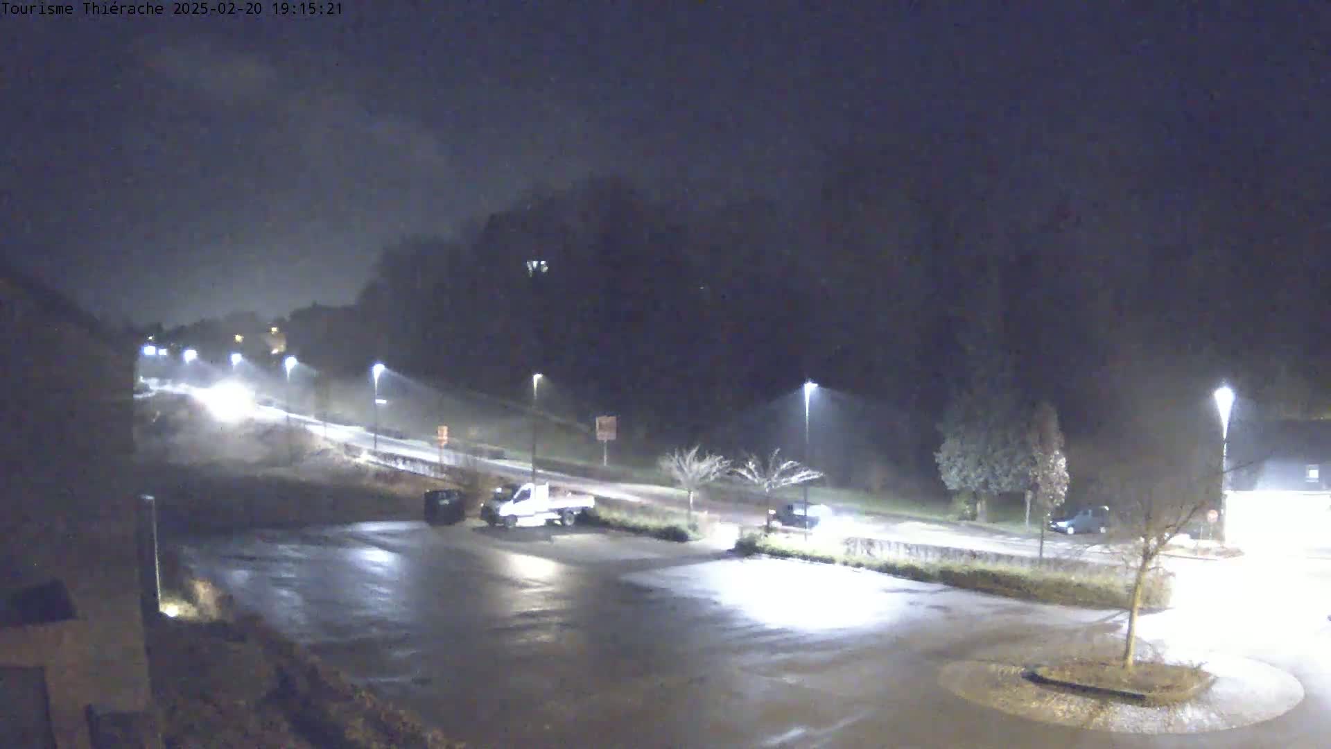 A nighttime view of a wet, empty parking lot and a road with a few vehicles, under streetlights, with a dark, tree-lined hill in the background.