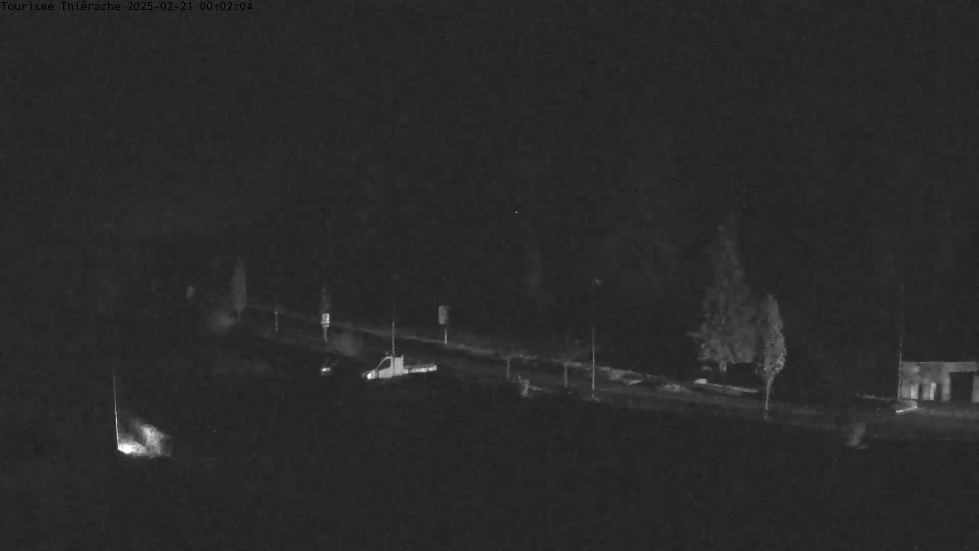 A nighttime, low-light image shows a road with a few vehicles and some trees under a dark sky.