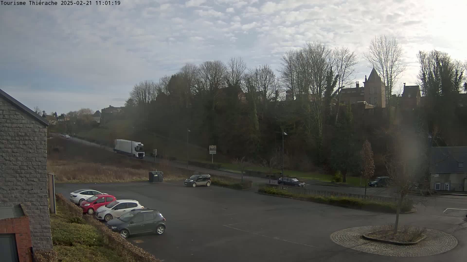 A mostly cloudy day reveals a parking lot with several cars, a large truck on a road, and a hilltop castle in the distance.