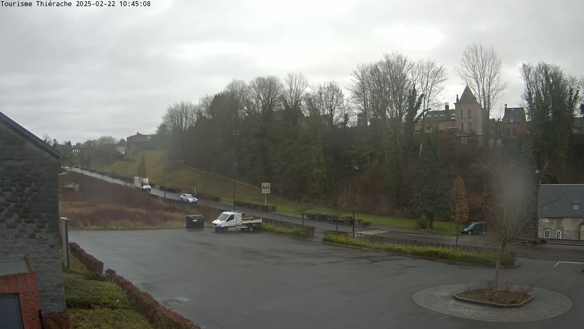 A mostly empty parking lot overlooks a road with several vehicles, leading to a hill topped with trees and buildings under an overcast sky.