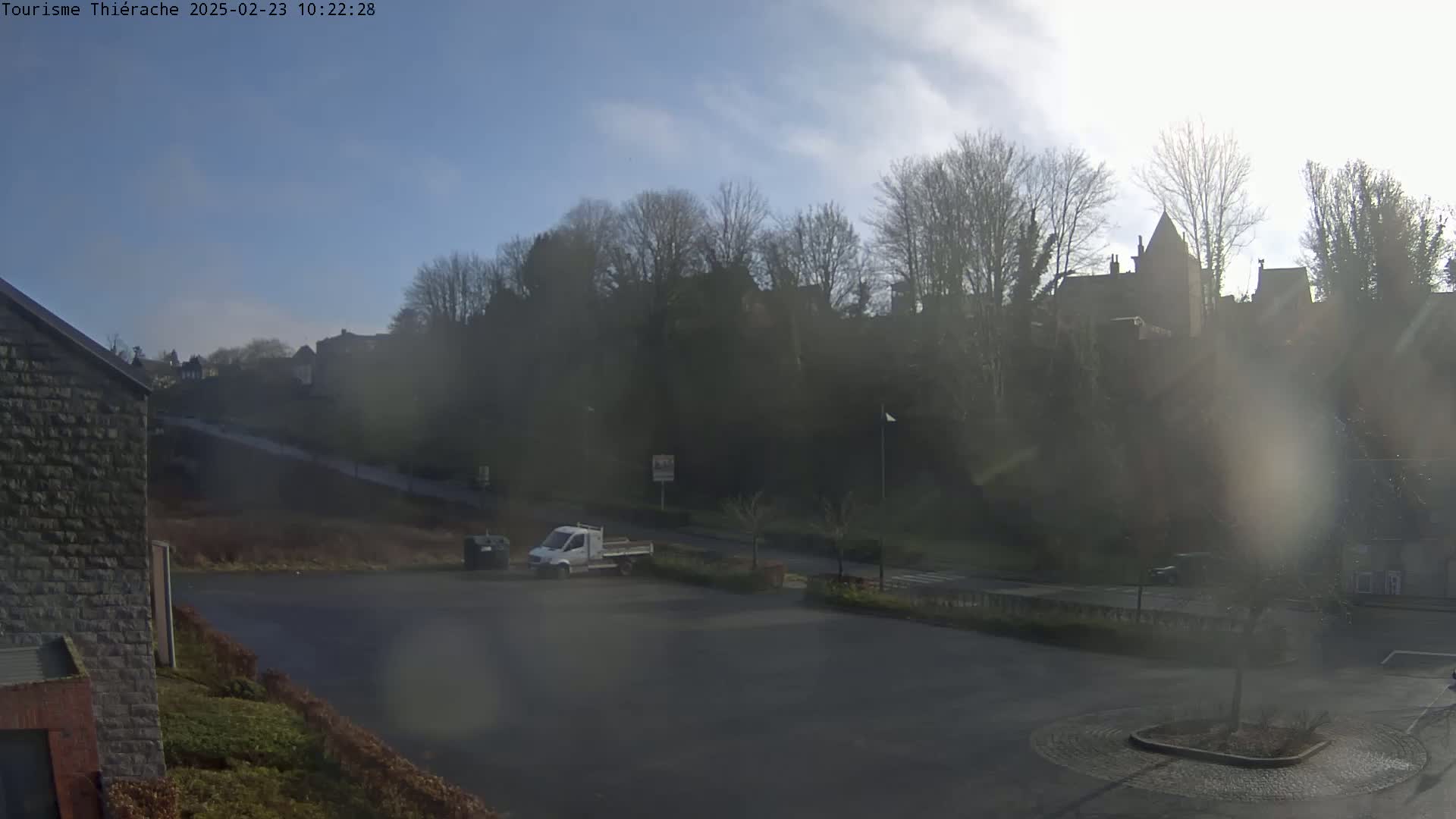 A hazy, sunny day reveals a parking lot with a white truck, a stone building, and a distant hill covered in leafless trees and buildings.
