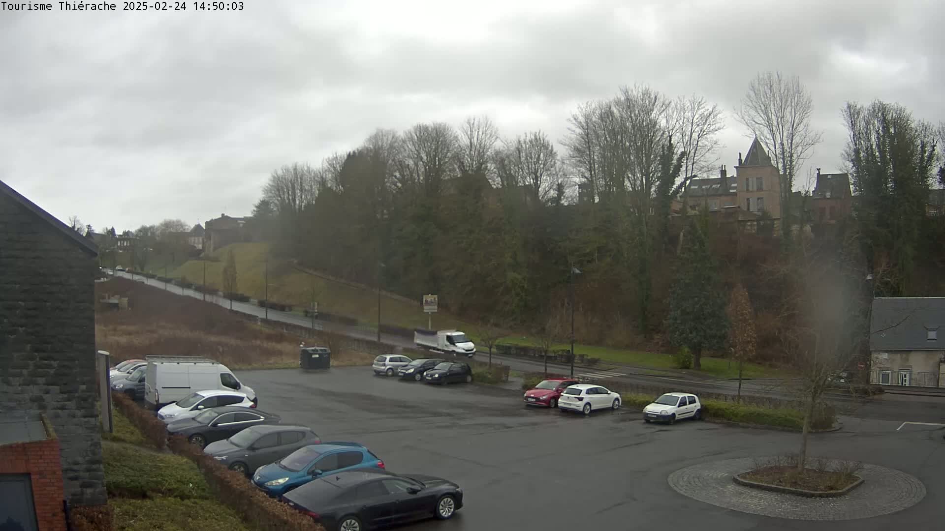 A parking lot with several cars and vans is situated below a hill topped with buildings and trees under an overcast sky.