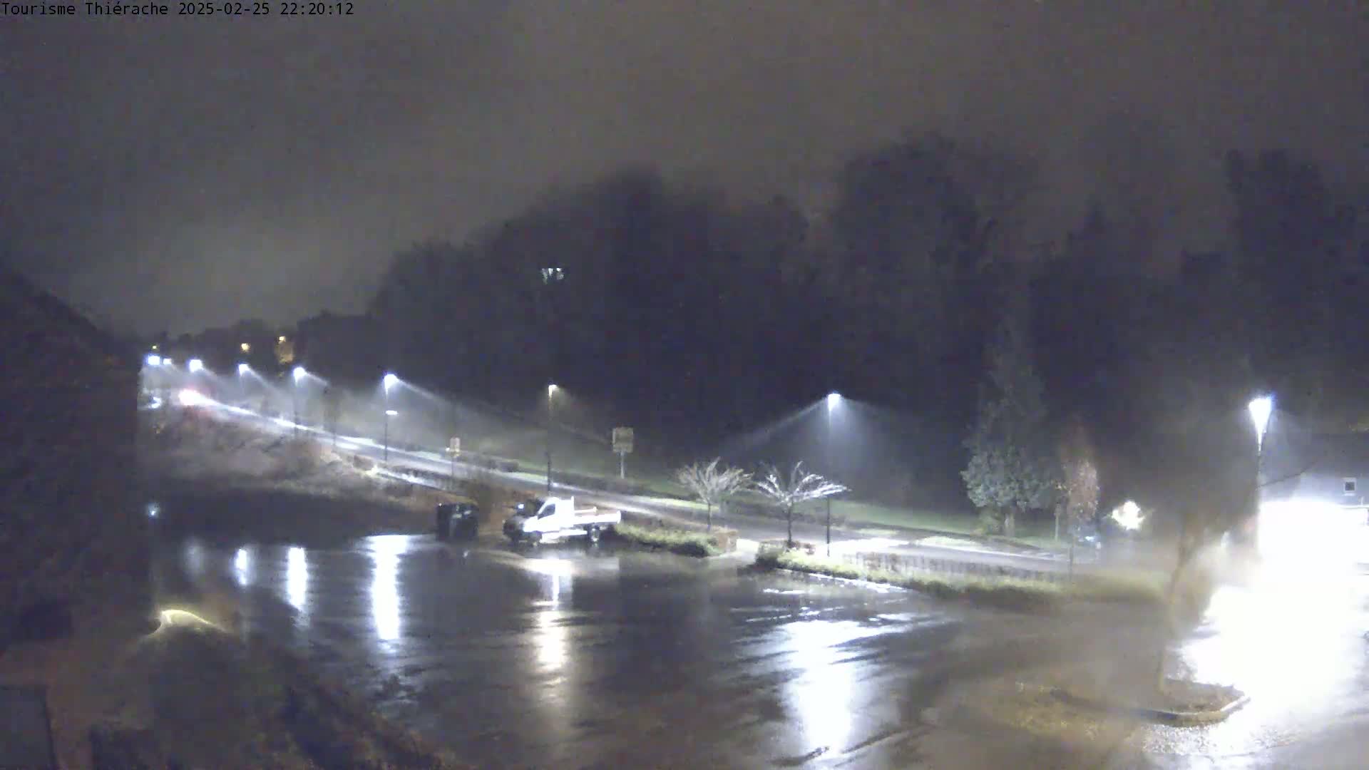 A wet, nighttime parking lot and road are illuminated by streetlights, with vehicles parked and driving under a dark, overcast sky.