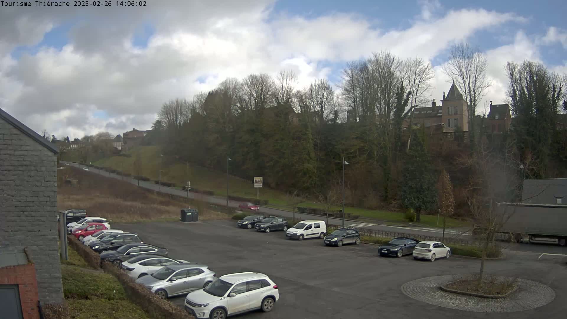 A parking lot filled with cars sits below a hill covered in bare trees, with a building and other structures visible in the distance under a partly cloudy sky.