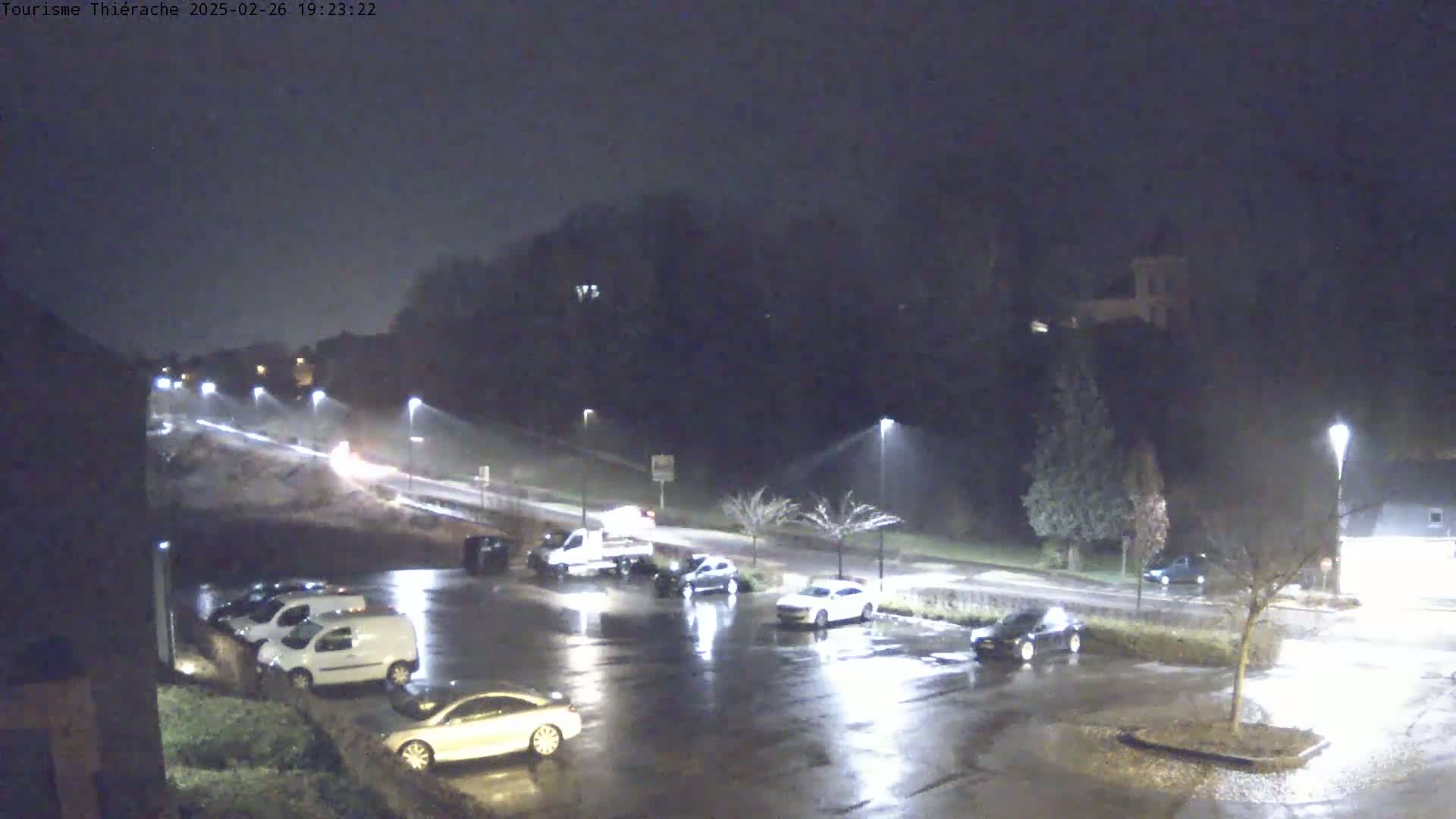 A nighttime, rain-slicked parking lot and road are illuminated by streetlights, showing several cars parked and a few driving by.