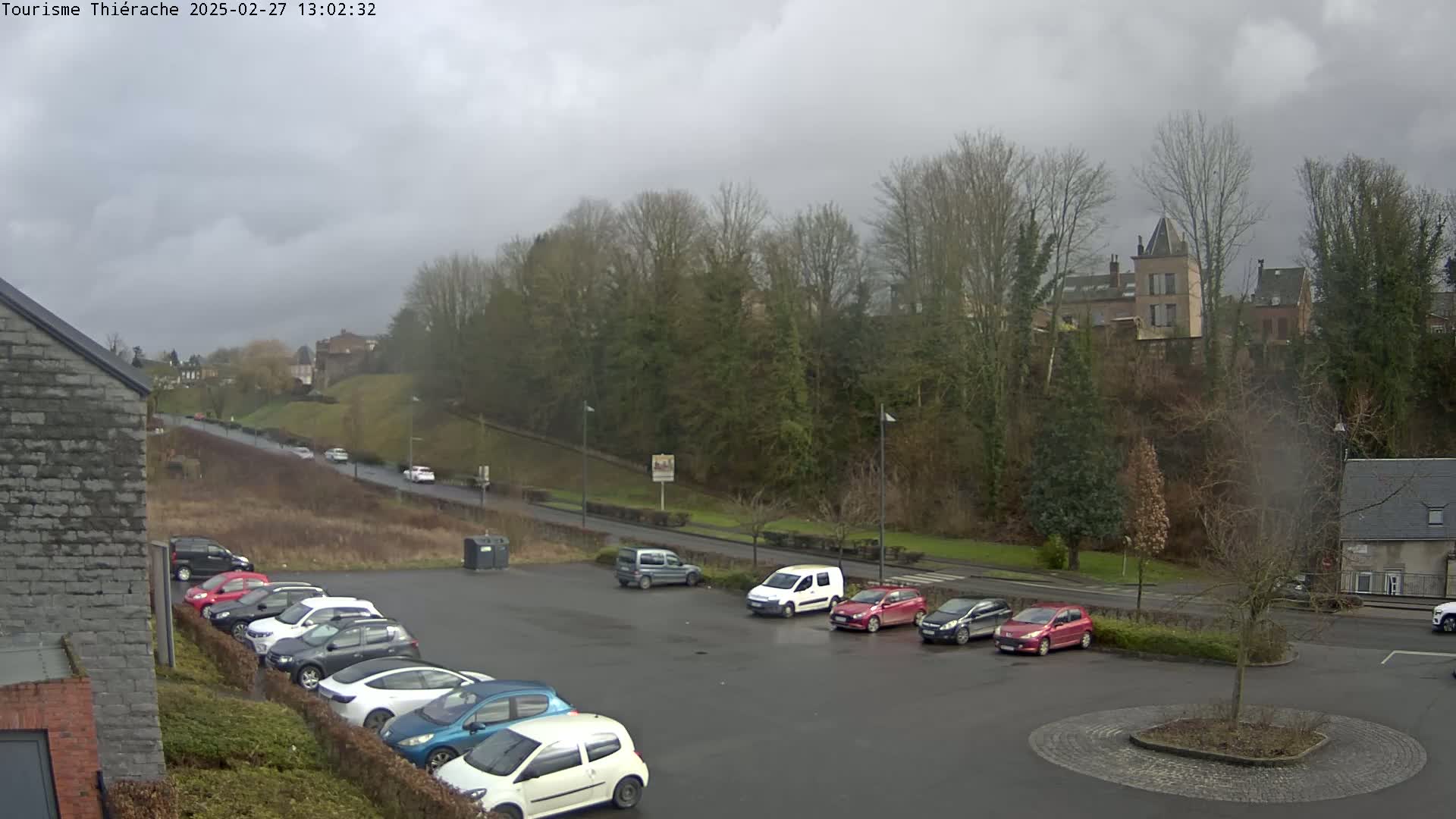 A parking lot with numerous cars is situated below a tree-covered hill on which buildings stand under an overcast sky.