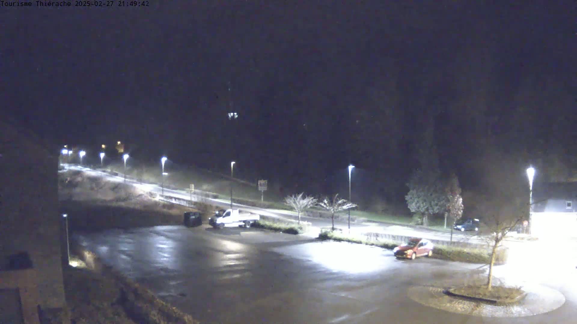 A nighttime view of a mostly empty parking lot with a few vehicles, illuminated by streetlights, next to a road that curves gently uphill.