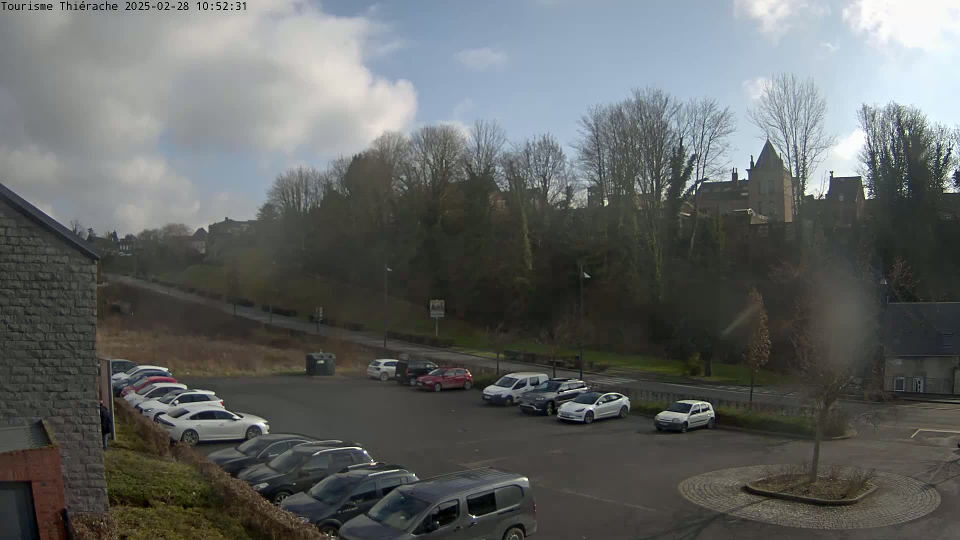 A parking lot with several cars is situated below a hill covered in leafless trees and buildings, under a partly sunny sky.