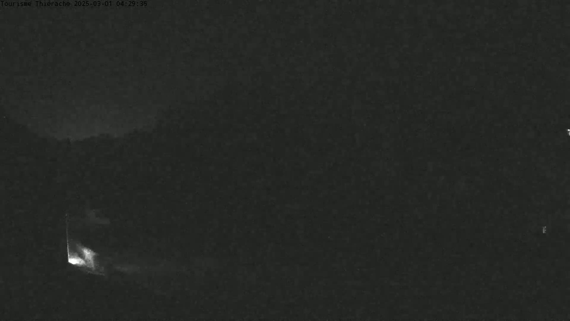 A low-light nighttime image shows a dark, possibly wooded area with a small bright light source emanating from what appears to be an opening or doorway.
