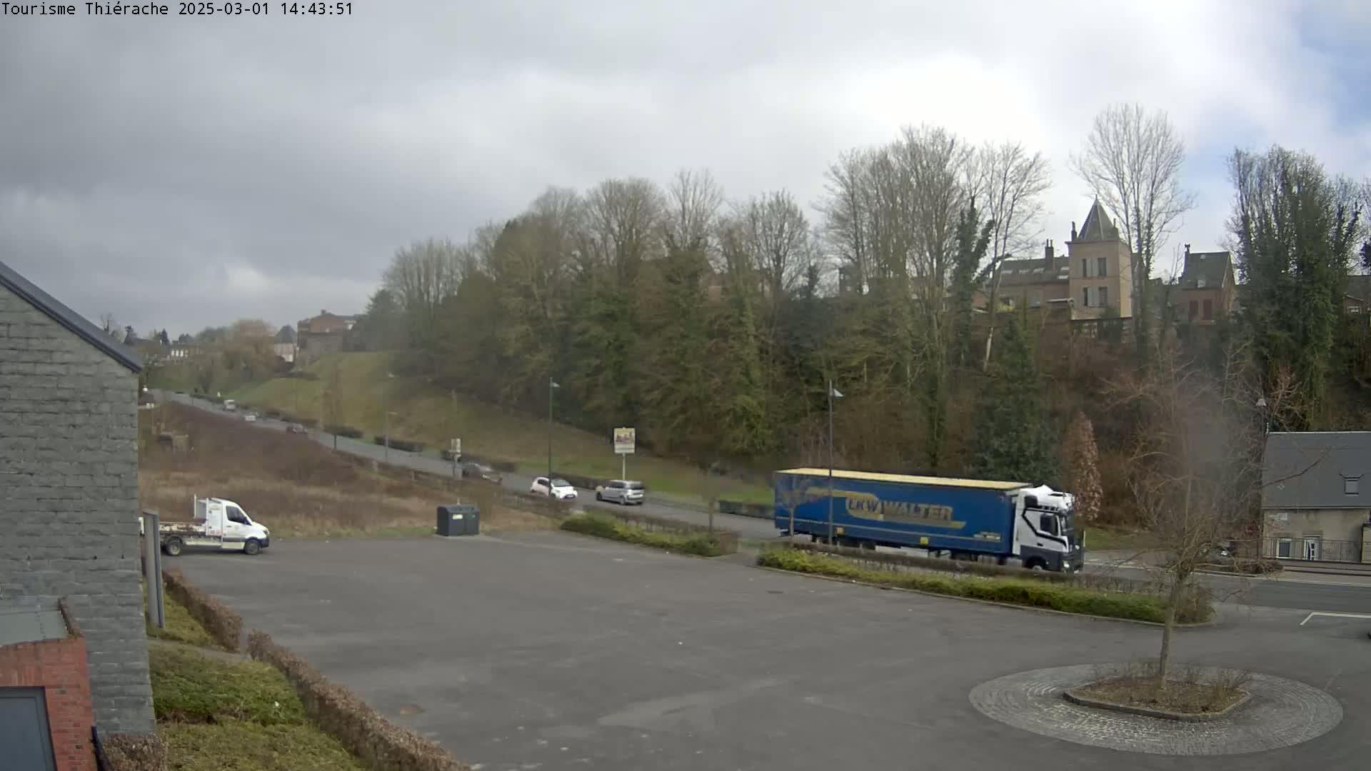 A mostly cloudy day reveals a semi-truck and several cars driving on a road that curves past a hill covered in bare trees and buildings.