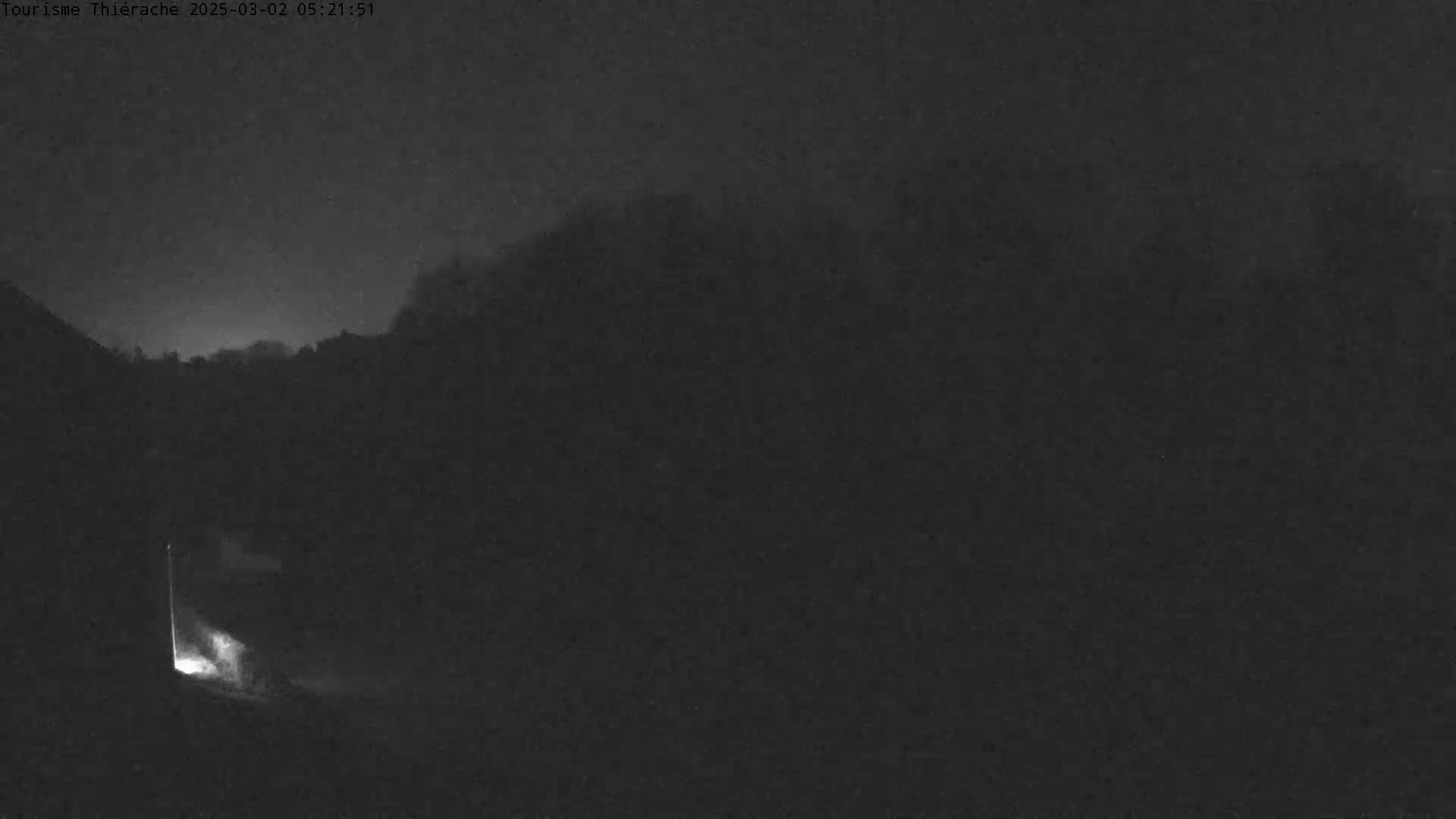 A nighttime, low-light image shows a dark, blurry landscape with a faint light source emanating from a structure in the bottom left corner.