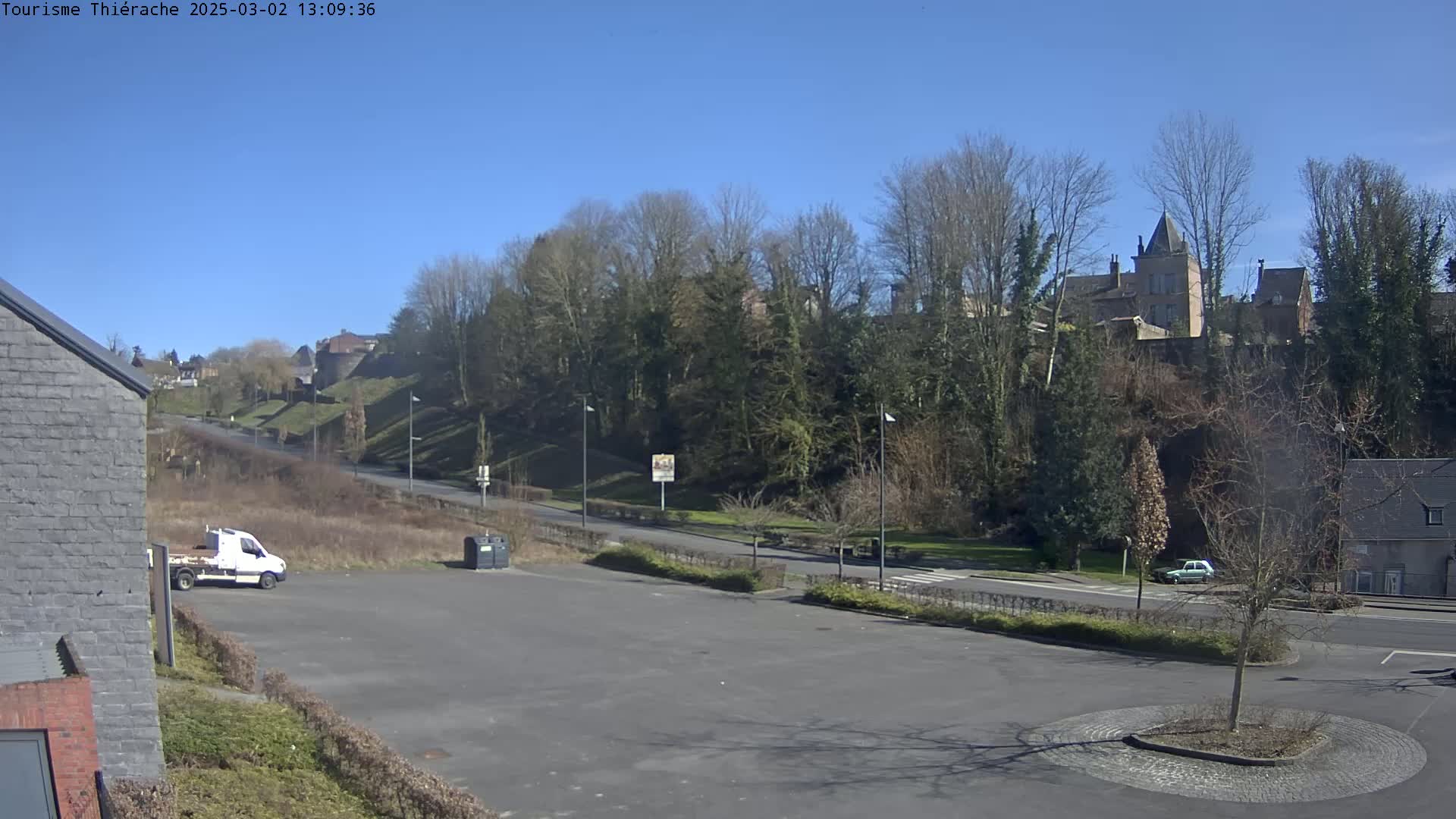 A mostly empty parking lot overlooks a road leading to a hill covered in leafless trees and buildings under a clear blue sky.