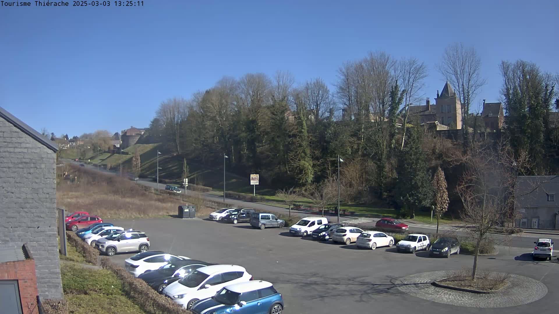 A parking lot filled with cars sits below a tree-covered hill on which a stone building is visible, all under a clear blue sky.