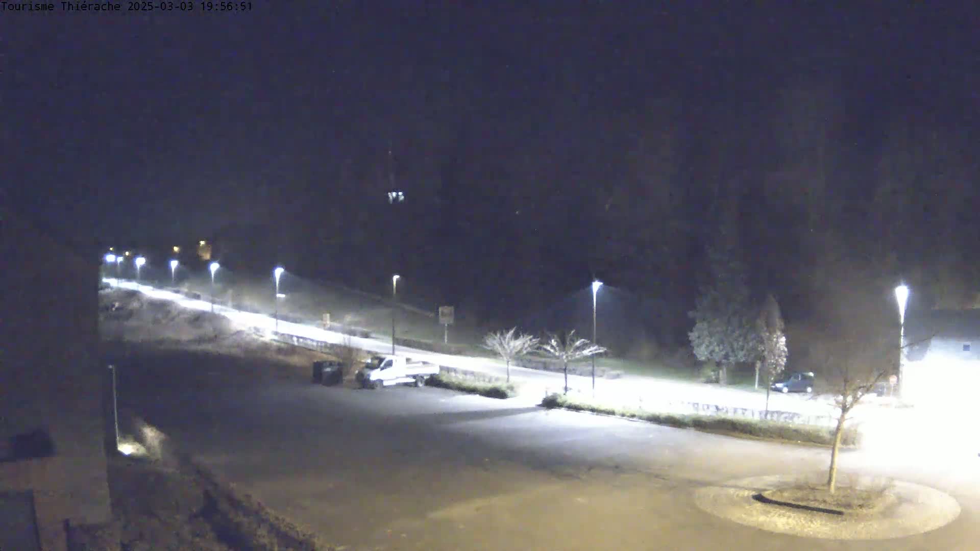 A nighttime view of a mostly empty parking lot and road, illuminated by streetlights, with a few vehicles present.