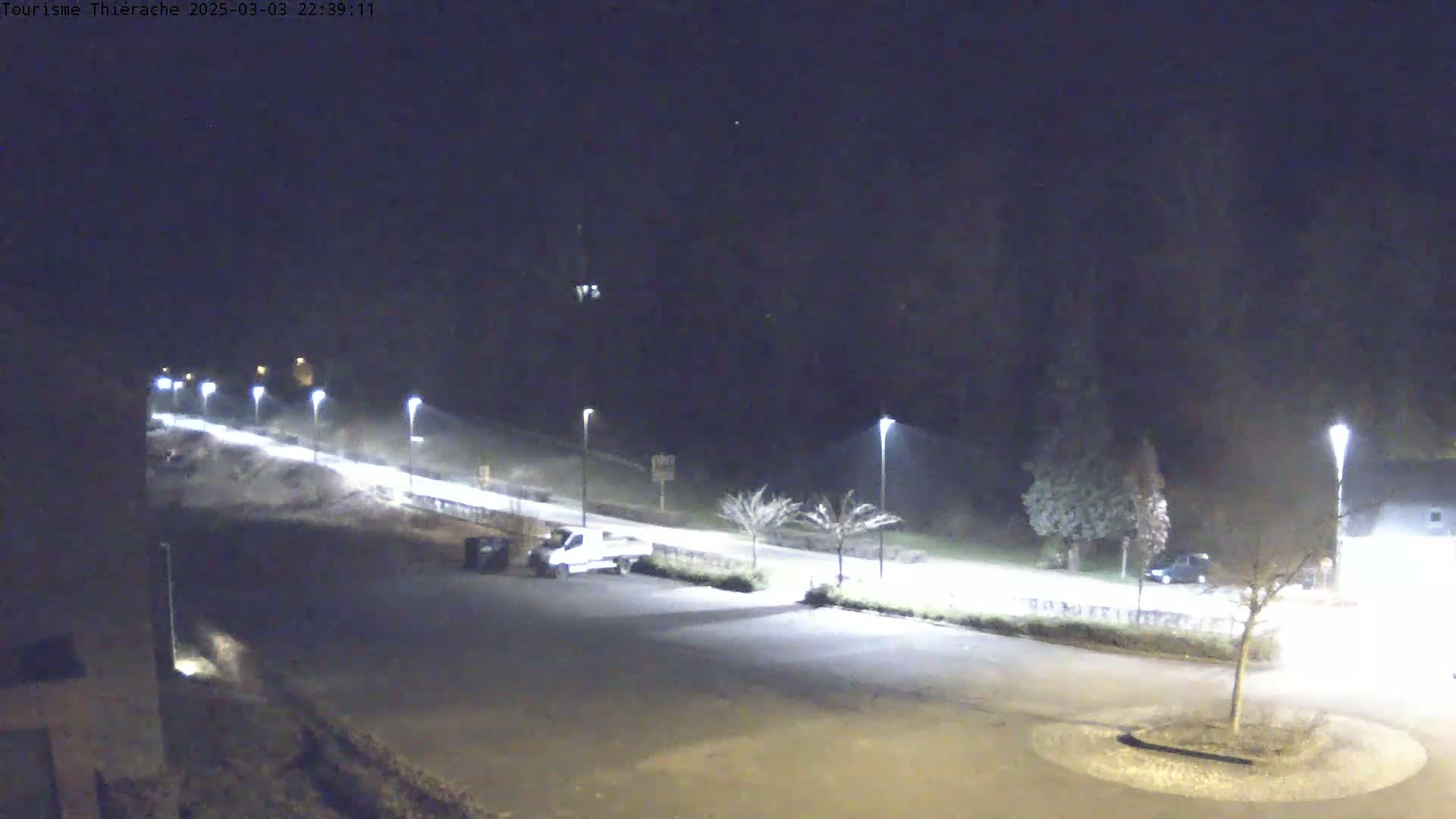 A nighttime, dimly lit parking lot and road scene with a few vehicles, streetlights, and bare trees under a clear, dark sky.
