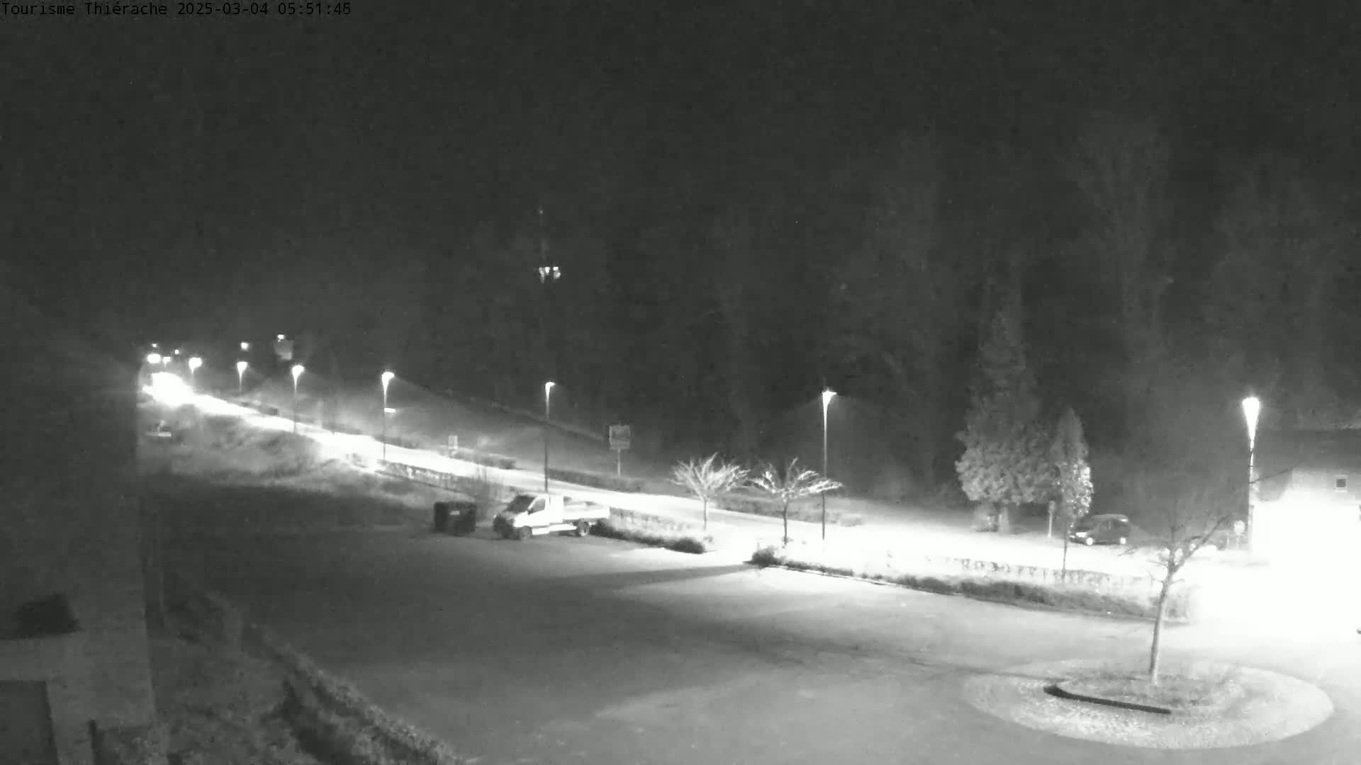 A nighttime, snow-covered parking lot and road are illuminated by streetlights, with a few vehicles visible.
