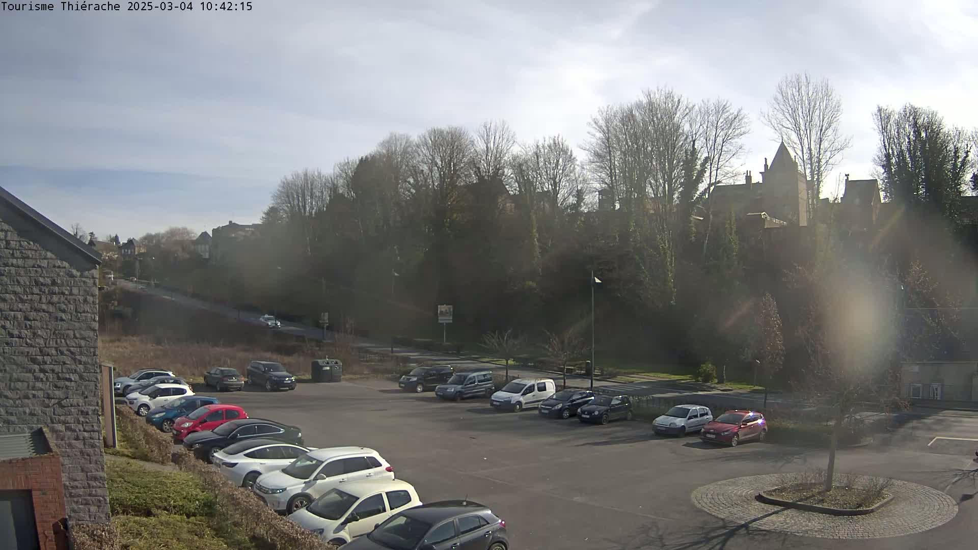 A parking lot with several cars is situated below a hill covered in bare trees and buildings under a partly sunny sky.