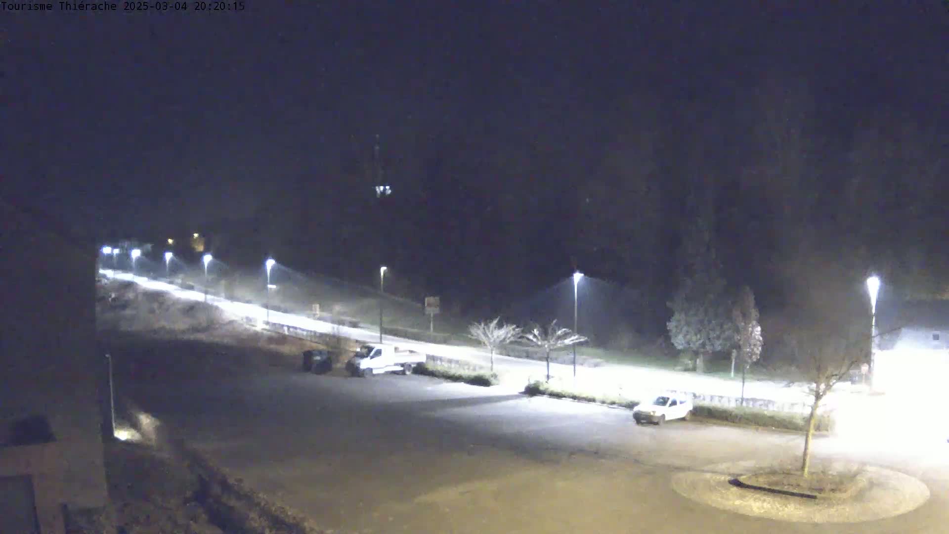 A nighttime view of a mostly empty parking lot and a road illuminated by streetlights, with light snow or rain falling.