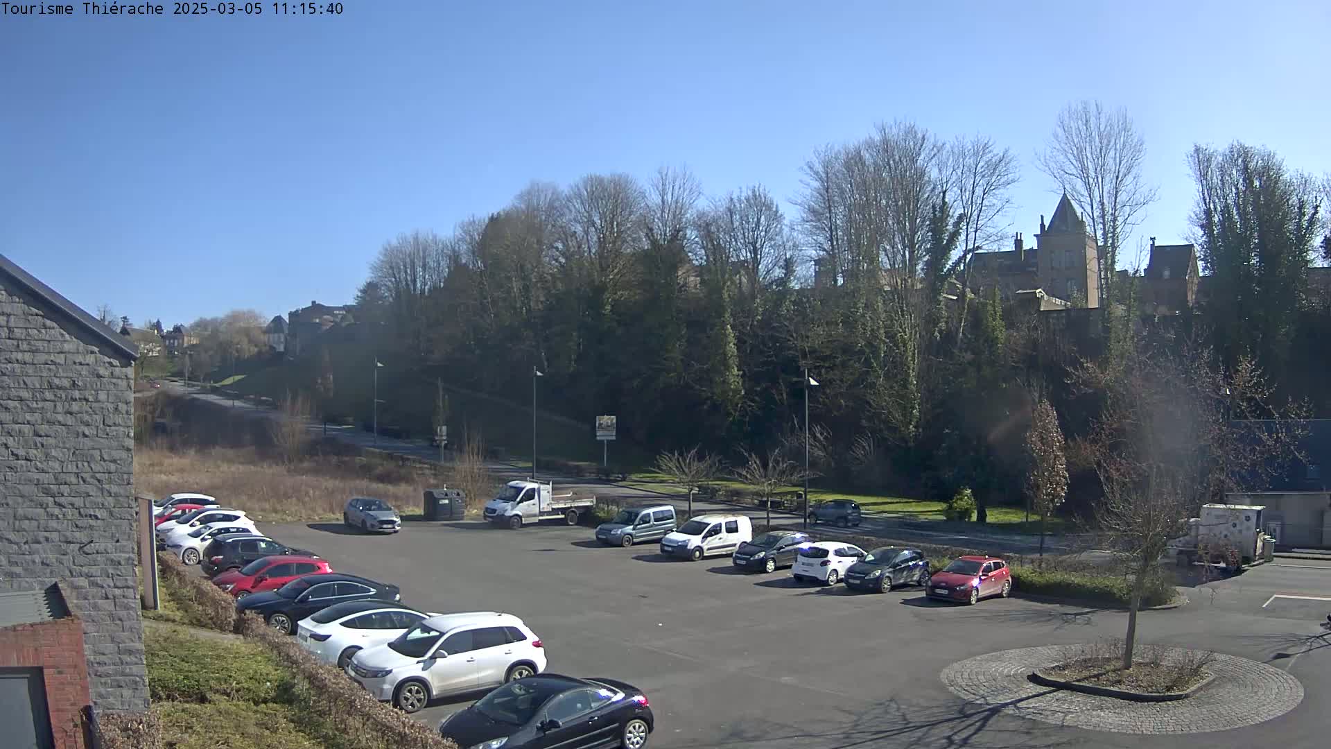 A parking lot filled with cars sits in front of a line of trees and buildings under a clear, sunny sky.