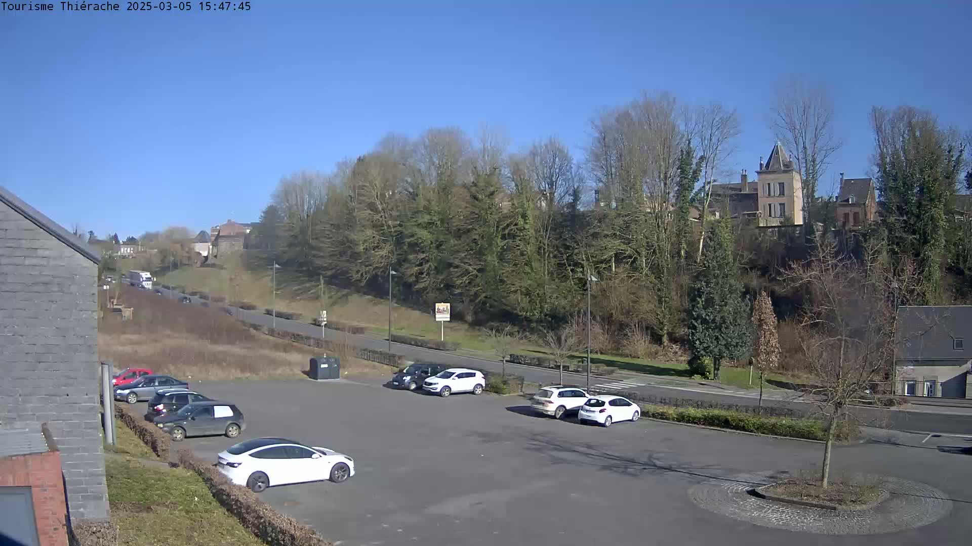 A sunny day reveals a parking lot with several cars, a road with sparse traffic, and a hillside with trees and buildings in the background.