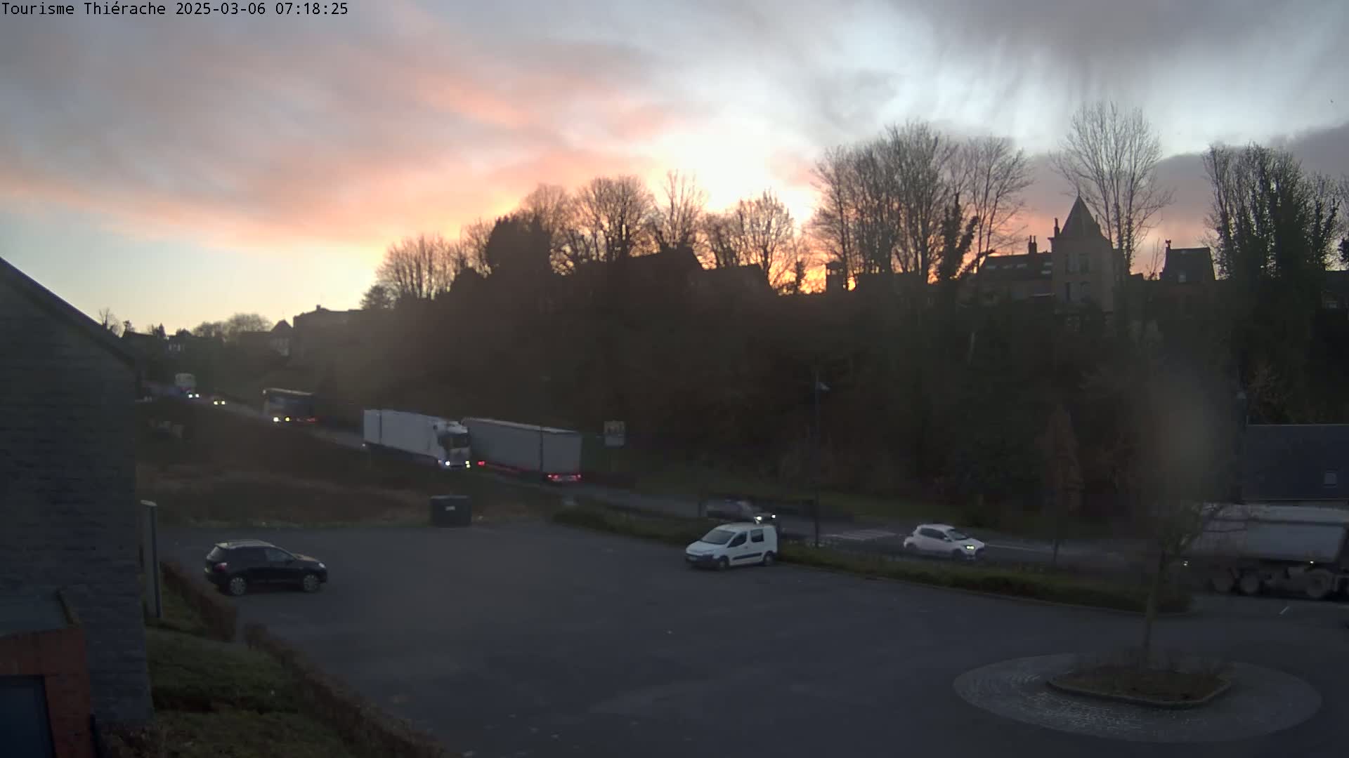 Several cars and trucks are parked and driving on a road near a hill with buildings and trees silhouetted against a dusky, pinkish-orange sunset.