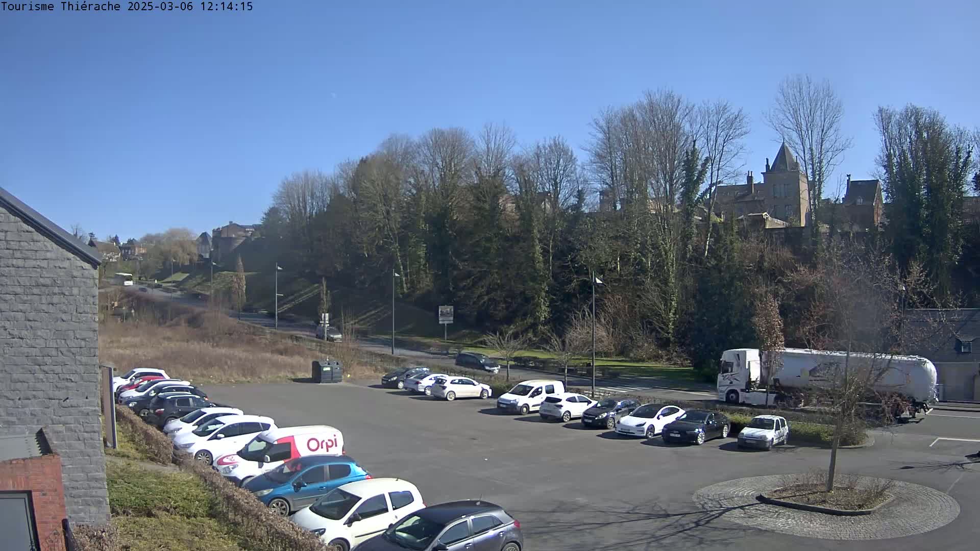 A sunny day reveals a parking lot full of cars near a road leading to a hill topped with trees and buildings.