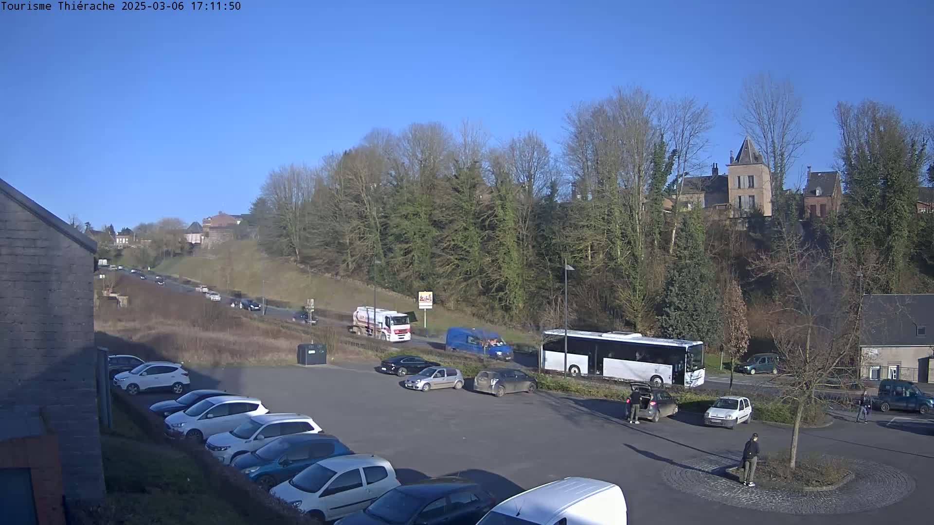 A parking lot with several cars is shown, next to a busy road with cars and trucks driving under a clear blue sky.