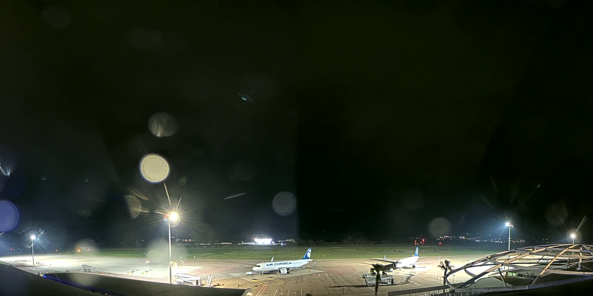 Two airplanes are parked on a brightly lit airport tarmac at night during a light rain.