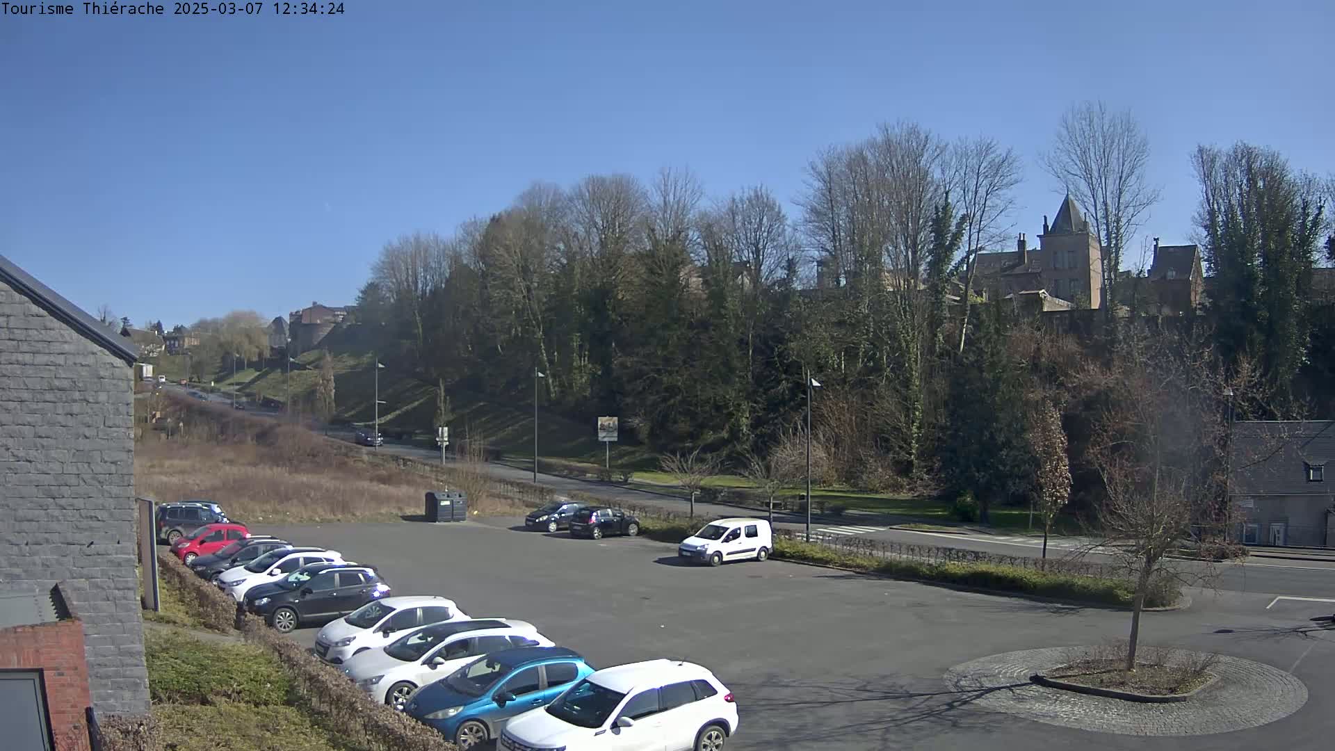 Under a clear blue sky, a parking lot filled with cars is situated near a tree-lined road leading to a distant hilltop castle.