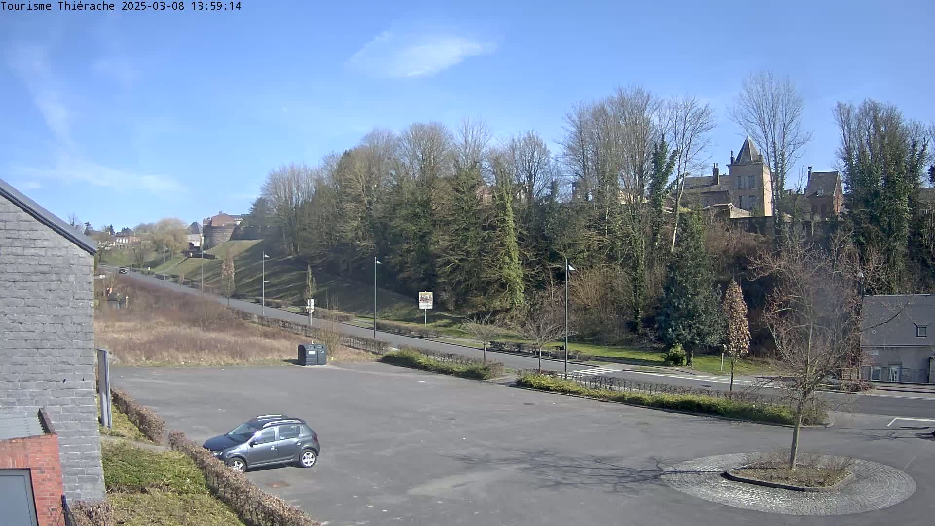 A mostly empty parking lot overlooks a road leading to a hill with trees and buildings, all under a clear blue sky.